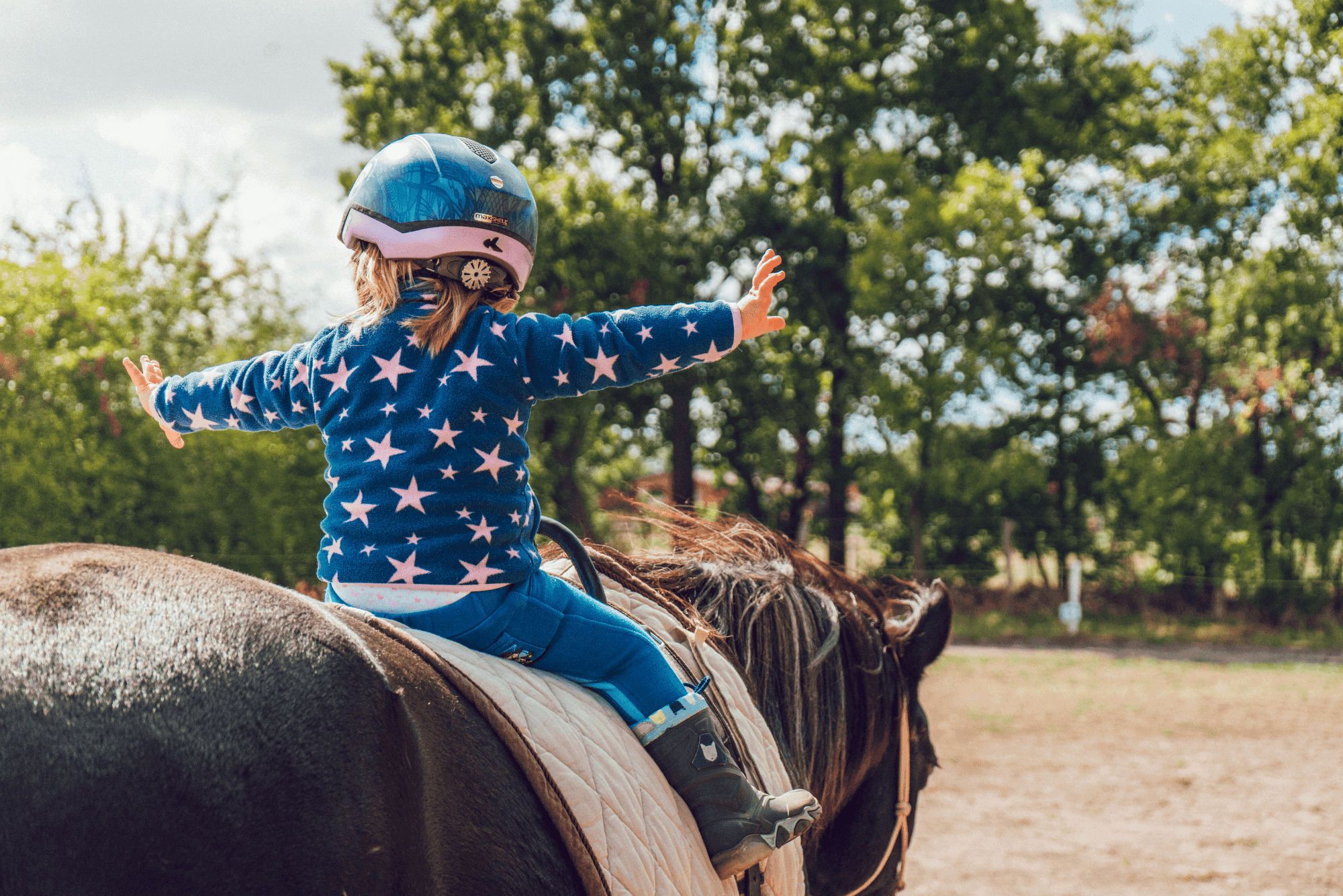 A little girl wearing blue and pink star-patterned and a helmet with stars on it is sitting in the saddle of a brown horse at an equestrian center for children riding horses, jumping around joyfully