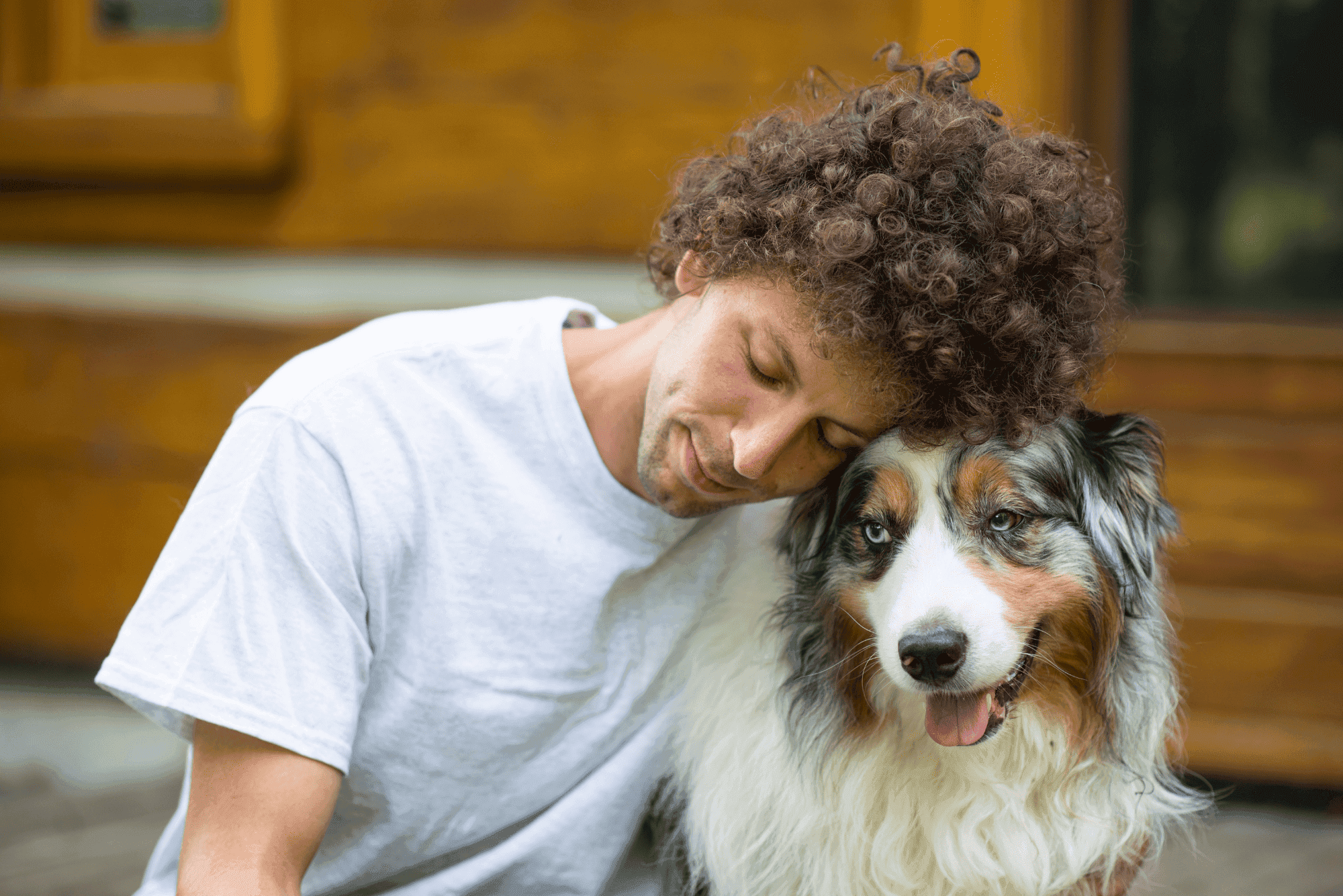 A young, curly-haired man in a white t-shirt hugging a happy Australian Shepherd dog